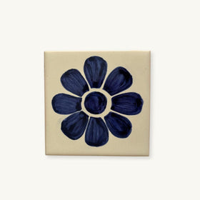 A square ceramic tile featuring a hand-painted dark blue flower motif on a warm cream background, shown against a plain off-white backdrop.
